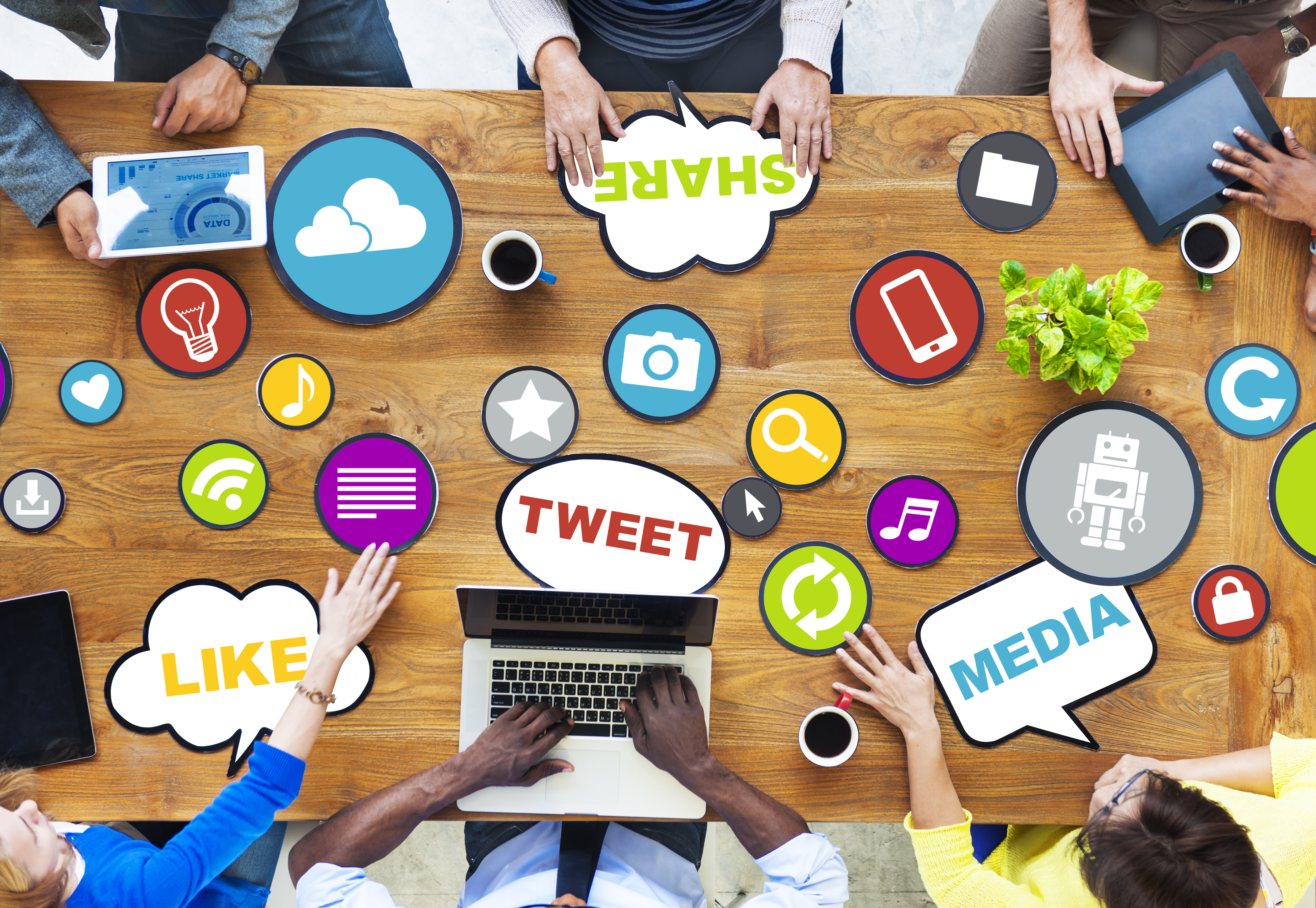 Multiple individuals seated around table with coffee and laptops brainstorming ideas with cutouts of words including: share, like, tweet, media